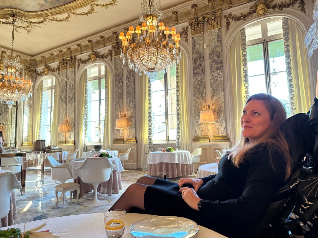 me (blonde woman with long hair dressed in black in a black wheelchair) in a large, ornate 1770's style dining room done in shades of cream and gold.

